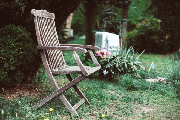 An image of a cemetery. In the foreground is an empty weathered wooden chair. Behind the chair is a headstone with a flowering plant in front of it. The grass is sparse.